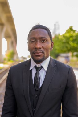 Portrait of handsome bearded African businessman in suit waiting at the railway train station outdoors