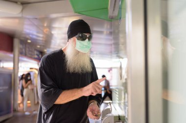 Portrait of mature bearded hipster man with mask for protection from corona virus outbreak social distancing at the skytrain station