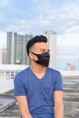 Portrait of young multi ethnic man with mask for protection from corona virus outbreak against view of the city at rooftop of building