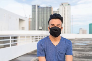 Portrait of young multi ethnic man with mask for protection from corona virus outbreak against view of the city at rooftop of building