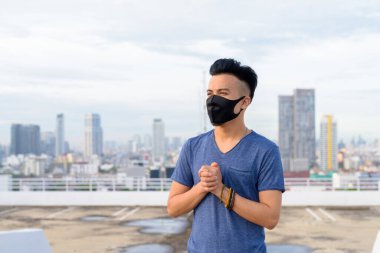 Portrait of young multi ethnic man with mask for protection from corona virus outbreak against view of the city at rooftop of building