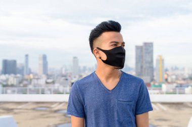 Portrait of young multi ethnic man with mask for protection from corona virus outbreak against view of the city at rooftop of building