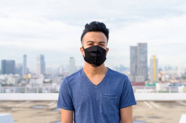 Portrait of young multi ethnic man with mask for protection from corona virus outbreak against view of the city at rooftop of building