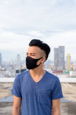 Portrait of young multi ethnic man with mask for protection from corona virus outbreak against view of the city at rooftop of building