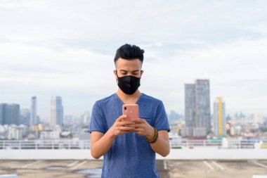 Portrait of young multi ethnic man with mask for protection from corona virus outbreak against view of the city at rooftop of building
