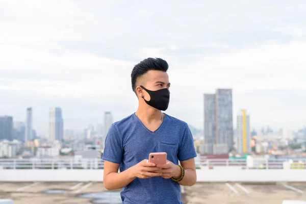 Portrait of young multi ethnic man with mask for protection from corona virus outbreak against view of the city at rooftop of building