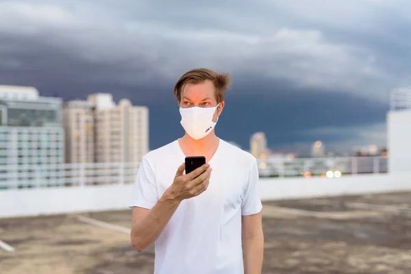 Portrait of young man with mask for protection from corona virus outbreak against view of the city during stormy weather outdoors