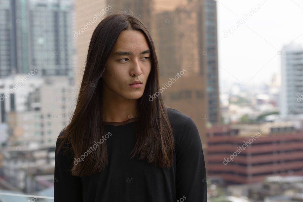 Portrait of young Asian man with long hair against view of the city outdoors