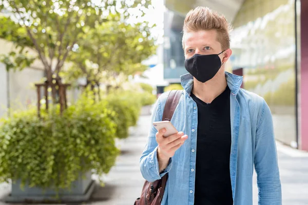 Portrait of blond tourist man wearing mask for protection from corona virus outbreak in the city streets with nature outdoors