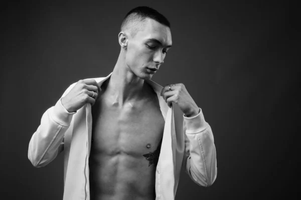 Studio shot of young handsome androgynous man with jacket fully unbuttoned against gray background in black and white