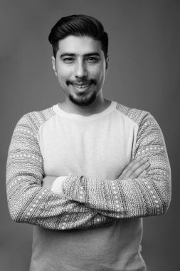 Siyah-beyaz gri arka planda günlük giysiler giyen genç sakallı İranlı adamın stüdyo fotoğrafı.