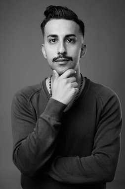 Bıyıklı, uzun kollu, gri arka planlı, siyah beyaz saçlı, yakışıklı İranlı genç adamın stüdyo fotoğrafı.