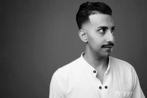 Studio shot of young handsome Iranian man with mustache wearing white shirt against gray background in black and white