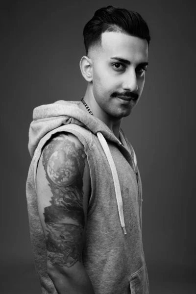 Studio shot of young handsome Iranian man with mustache wearing hooded shirt against gray background in black and white