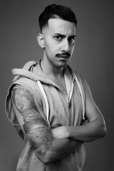 Studio shot of young handsome Iranian man with mustache wearing hooded shirt against gray background in black and white