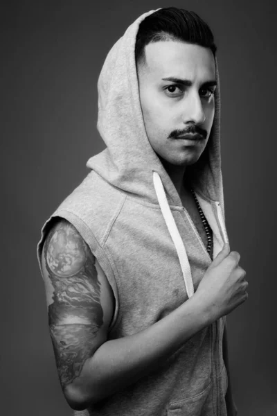 Studio shot of young handsome Iranian man with mustache wearing hooded shirt against gray background in black and white