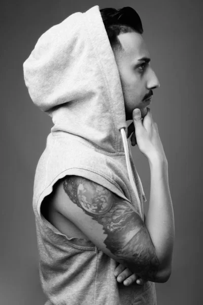 Studio shot of young handsome Iranian man with mustache wearing hooded shirt against gray background in black and white