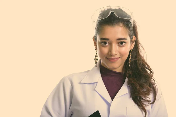 Studio shot of young beautiful Indian woman doctor as scientist wearing protective glasses isolated against white background