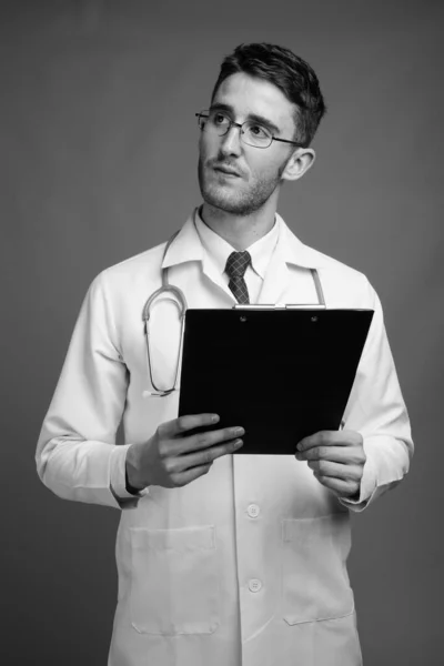 年轻英俊男医生拿着眼镜 拿着剪贴板 背景灰暗 黑白相间的摄影棚照片 — 图库照片