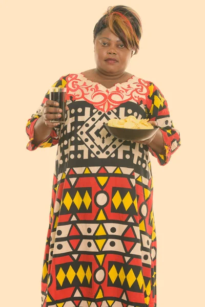 Photo de Studio de grosse femme africaine noire permanent tout en tenant le bol de croustilles et de verre de boisson soda — Photo