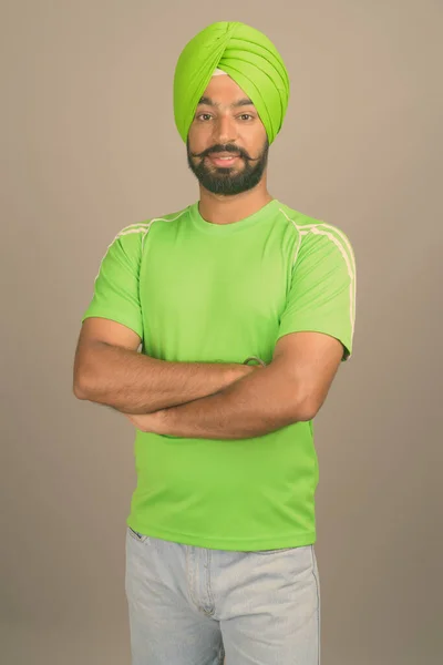 Młody przystojny indyjski Sikh człowiek noszenie turban na szarym tle — Zdjęcie stockowe