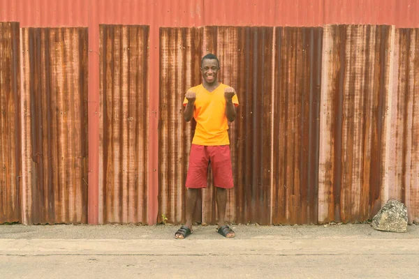 Νεαρός ευτυχισμένος μαύρος Αφρικανός που χαμογελάει και δείχνει ενθουσιασμένος ενώ στέκεται απέναντι στον παλιό σκουριασμένο τοίχο. — Φωτογραφία Αρχείου