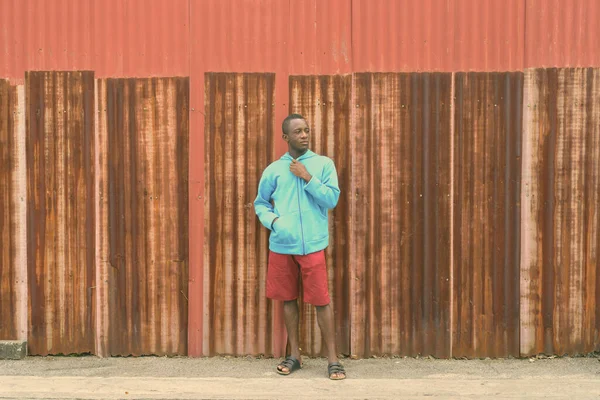 Νεαρός μαύρος Αφρικανός στέκεται ενώ σκέφτεται και κρατά το σακάκι ενάντια στο παλιό σκουριασμένο τοίχο φύλλων — Φωτογραφία Αρχείου