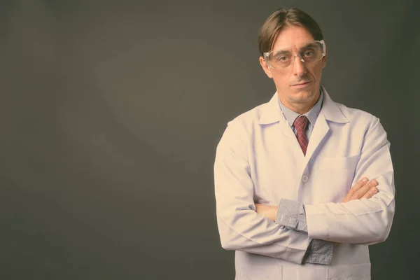 Mature handsome Italian man doctor wearing protective glasses against gray background — Stock Photo, Image