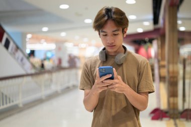 20 yaşında boynunda kulaklıkla Taylandlı bir adam alışveriş merkezinin koridorunda telefonuna gülümserken, mesajlaşırken, online alışveriş yaparken ve şehir yaşamında.