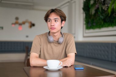 Kahverengi tişörtlü genç Taylandlı öğrenci sosyal medya, uzaktan iletişim ve dijital yaşam tarzı gibi modern bir kafede boynuna kulaklık takıyor..