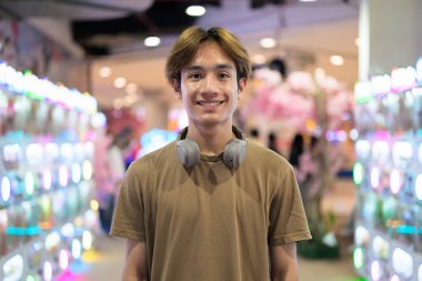 Kahverengi tişörtlü, kulaklıklı Taylandlı genç bir adam. Parlak bir alışveriş merkezinde, modern yaşam tarzında, teknoloji ve eğlence konseptinde otomatik satış makinesi kullanmaya hazır..