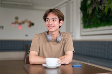 Kahverengi tişörtlü genç Taylandlı öğrenci sosyal medya, uzaktan iletişim ve dijital yaşam tarzı gibi modern bir kafede boynuna kulaklık takıyor..