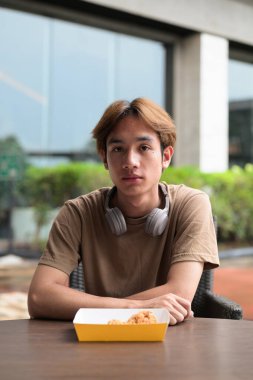 Fast food, aperatif ve yemek konsepti için açık bir masada kızarmış tavuk budu yemeye hazır kulaklıklı, kahverengi tişörtlü genç Asyalı öğrenci.