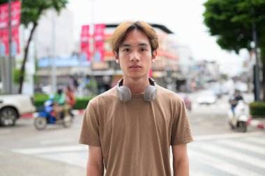 Kulaklıklı yakışıklı Asyalı genç Taylandlı yaz boyunca şehirde modern hayatın tadını çıkarıyor ve kameraya bakıyor.