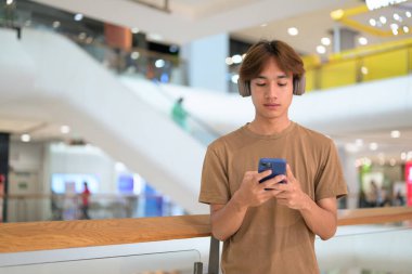 20 yaşında boynunda kulaklıkla Taylandlı bir adam alışveriş merkezinin koridorunda telefonuna gülümserken, mesajlaşırken, online alışveriş yaparken ve şehir yaşamında.