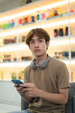 Kahverengi tişörtlü genç Taylandlı öğrenci sosyal medya, uzaktan iletişim ve dijital yaşam tarzı gibi modern bir kafede boynuna kulaklık takıyor..