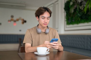 Kahverengi tişörtlü genç Taylandlı öğrenci sosyal medya, uzaktan iletişim ve dijital yaşam tarzı gibi modern bir kafede boynuna kulaklık takıyor..