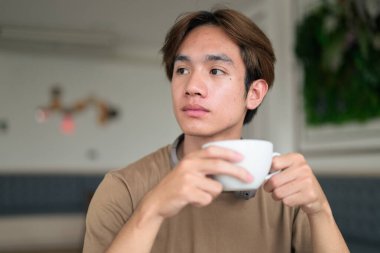 Kahverengi tişörtlü genç Taylandlı öğrenci sosyal medya, uzaktan iletişim ve dijital yaşam tarzı gibi modern bir kafede boynuna kulaklık takıyor..