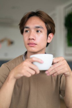 Kahverengi tişörtlü genç Taylandlı öğrenci sosyal medya, uzaktan iletişim ve dijital yaşam tarzı gibi modern bir kafede boynuna kulaklık takıyor..