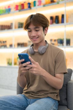 Kahverengi tişörtlü genç Taylandlı öğrenci sosyal medya, uzaktan iletişim ve dijital yaşam tarzı gibi modern bir kafede boynuna kulaklık takıyor..