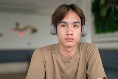 Kahverengi tişörtlü genç Taylandlı öğrenci sosyal medya, uzaktan iletişim ve dijital yaşam tarzı gibi modern bir kafede boynuna kulaklık takıyor..