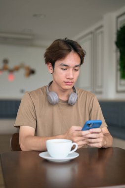 Kahverengi tişörtlü genç Taylandlı öğrenci sosyal medya, uzaktan iletişim ve dijital yaşam tarzı gibi modern bir kafede boynuna kulaklık takıyor..