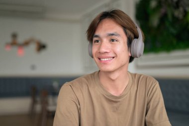 Kahverengi tişörtlü genç Taylandlı öğrenci sosyal medya, uzaktan iletişim ve dijital yaşam tarzı gibi modern bir kafede boynuna kulaklık takıyor..