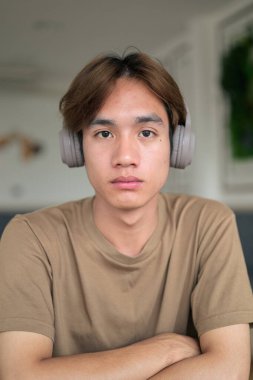 Kahverengi tişörtlü genç Taylandlı öğrenci sosyal medya, uzaktan iletişim ve dijital yaşam tarzı gibi modern bir kafede boynuna kulaklık takıyor..