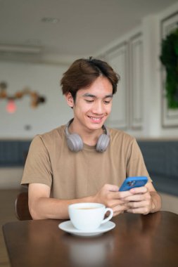 Kahverengi tişörtlü genç Taylandlı öğrenci sosyal medya, uzaktan iletişim ve dijital yaşam tarzı gibi modern bir kafede boynuna kulaklık takıyor..