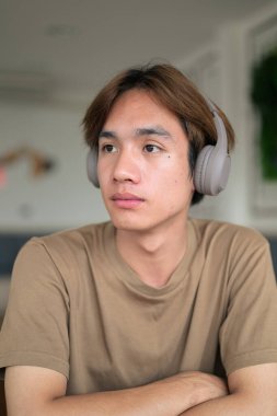 Kahverengi tişörtlü genç Taylandlı öğrenci sosyal medya, uzaktan iletişim ve dijital yaşam tarzı gibi modern bir kafede boynuna kulaklık takıyor..