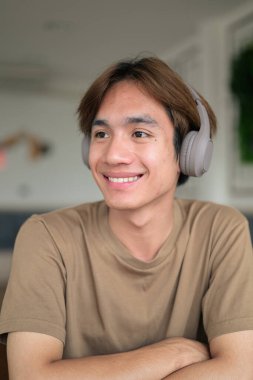 Kahverengi tişörtlü genç Taylandlı öğrenci sosyal medya, uzaktan iletişim ve dijital yaşam tarzı gibi modern bir kafede boynuna kulaklık takıyor..