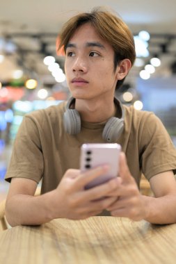 Kahverengi tişörtlü genç Taylandlı öğrenci sosyal medya, uzaktan iletişim ve dijital yaşam tarzı gibi modern bir kafede boynuna kulaklık takıyor..