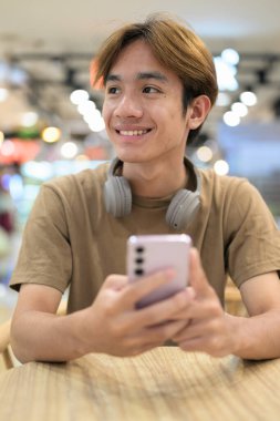 Kahverengi tişörtlü genç Taylandlı öğrenci sosyal medya, uzaktan iletişim ve dijital yaşam tarzı gibi modern bir kafede boynuna kulaklık takıyor..