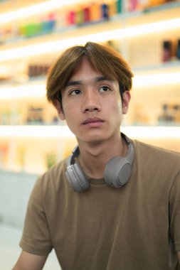 Kahverengi tişörtlü genç Taylandlı öğrenci sosyal medya, uzaktan iletişim ve dijital yaşam tarzı gibi modern bir kafede boynuna kulaklık takıyor..